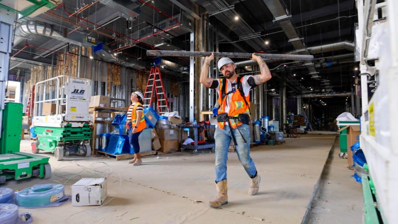 A construction worker carries a beam while wearing an exoskeleton for support. 